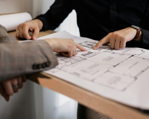 Two architects reviewing and pointing at a detailed floor plan on paper in an office setting.